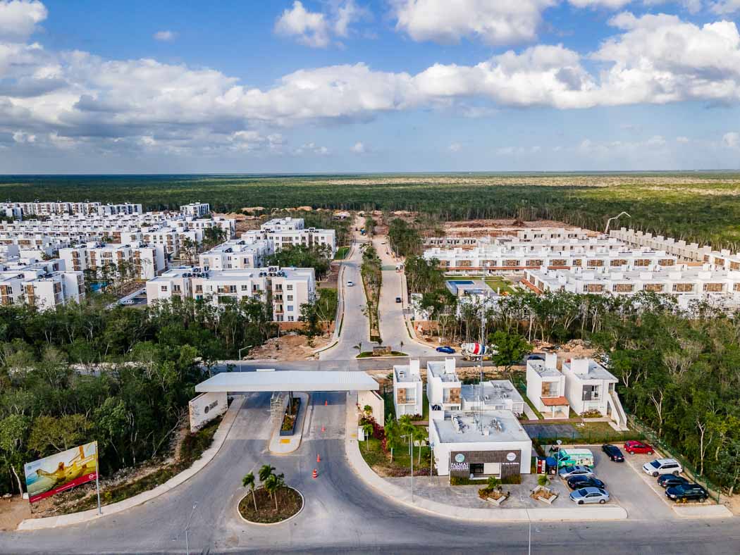 Visa completa del desarrollo Palmas Turquesa en la Riviera Maya. Carros estacionados a lado derecho de la entrada. Dentro hay casas y departamentos.