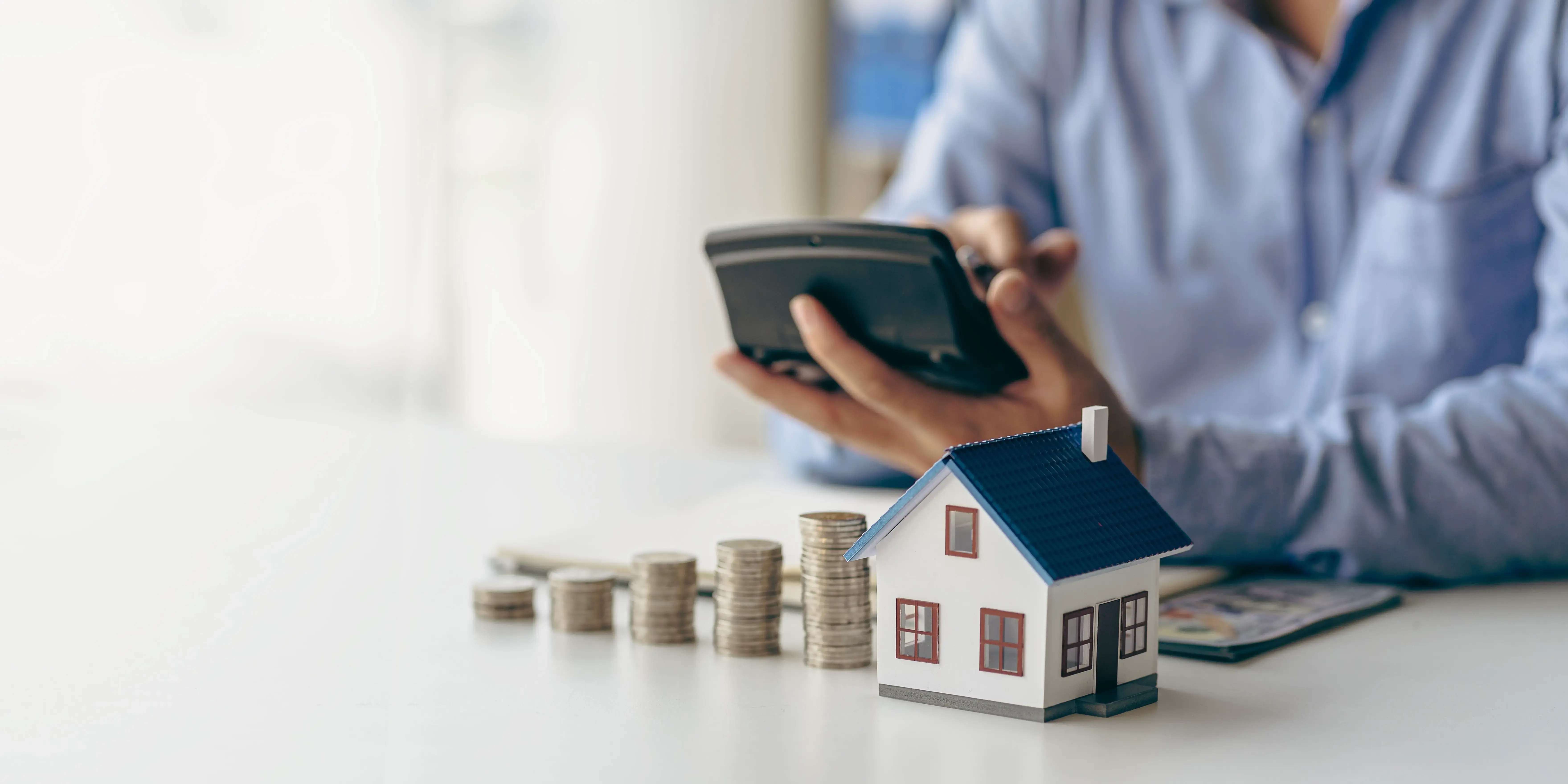 Persona sosteniendo una calculadora y haciendo cuentas frente unas torres de monedas y una casa de madera para el programa nivela tu pago Infonavit