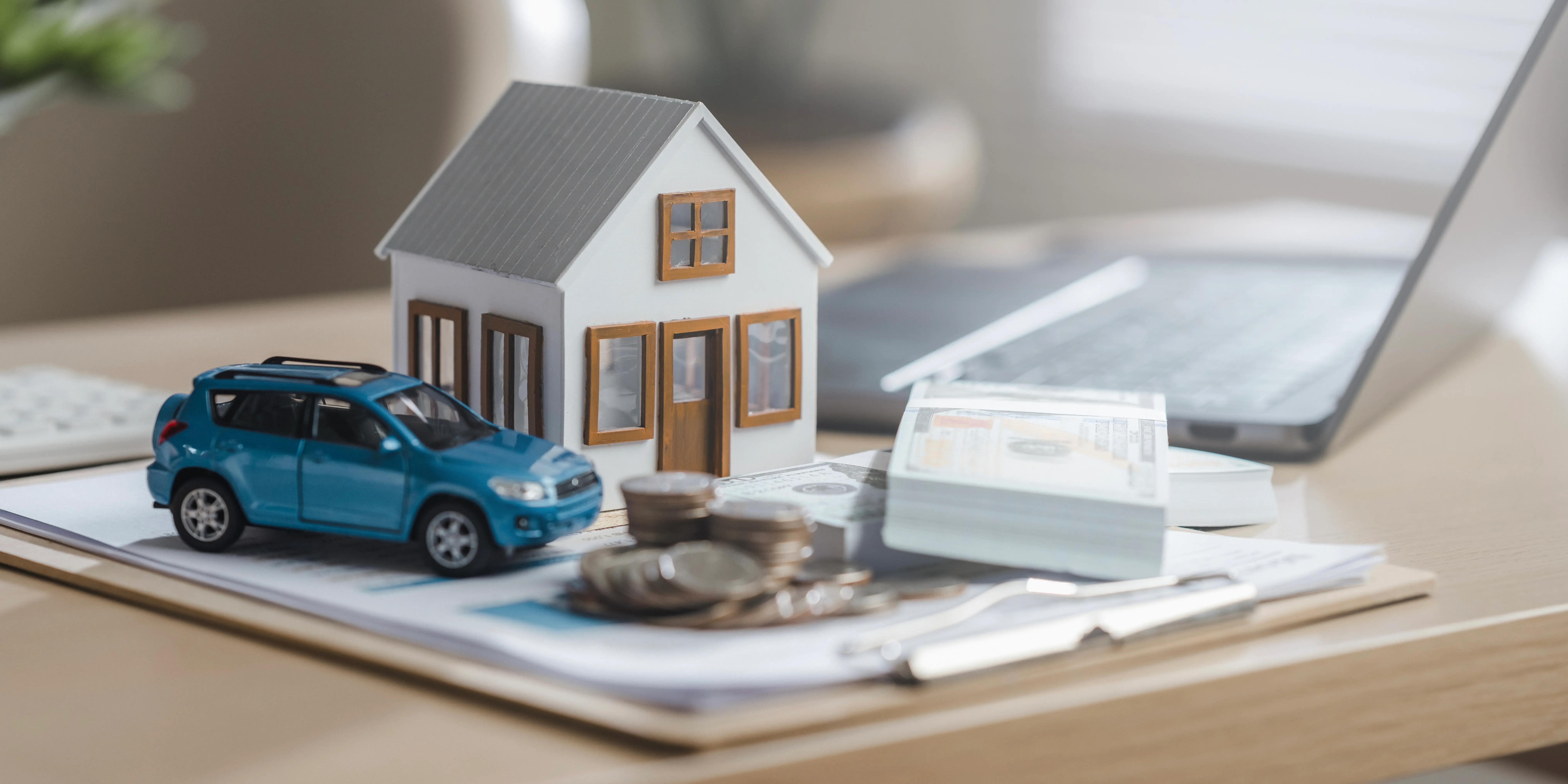 Un coche azul y una casa de juguete encima de una pila de monedas y a un lado una computadora para tener la planificación patrimonial y para inversión