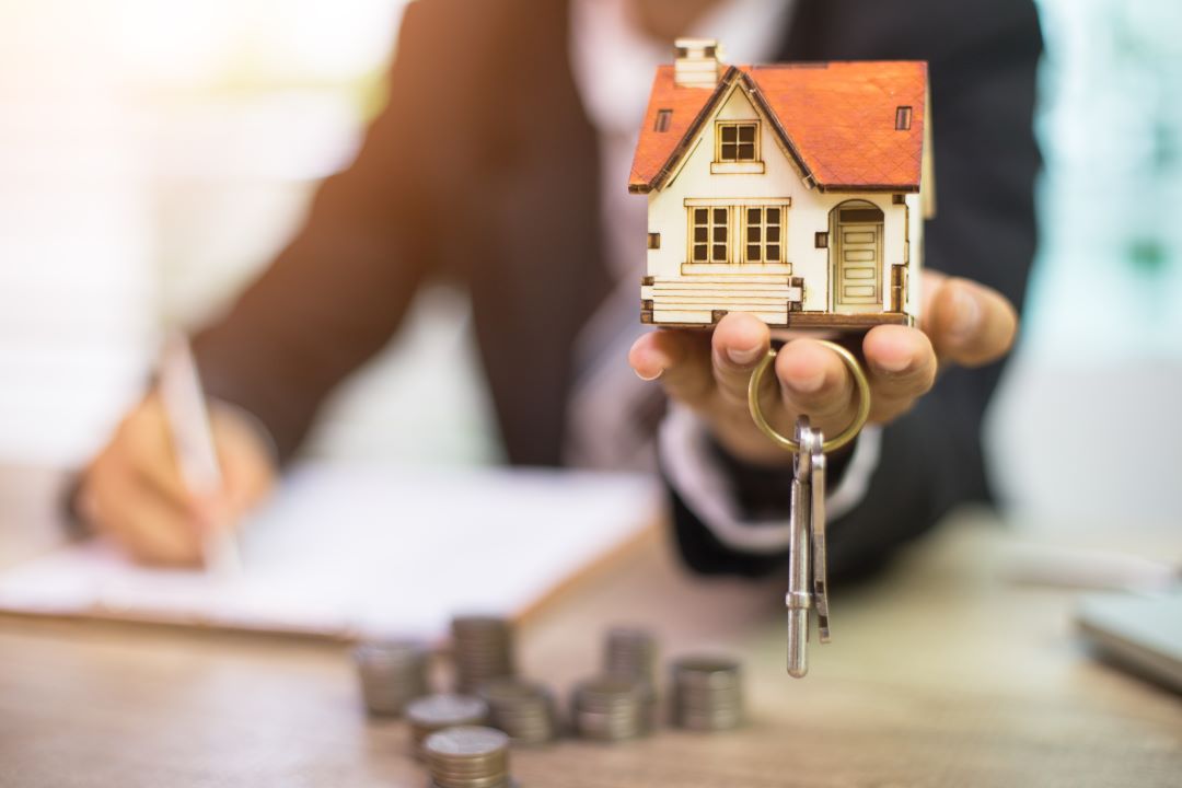 Una persona en su escritorio, donde hay monedas apiladas y un documento, extiende una casita con llaves para firmar un trato. 