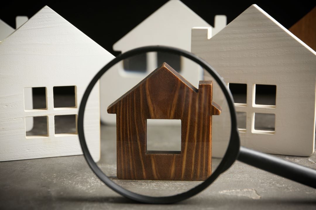 Figuras bidimensionales de casas, tres blancas al rededor de una color madera con betas. Hay una lupa enfocando a la color madera. Suelo gris.