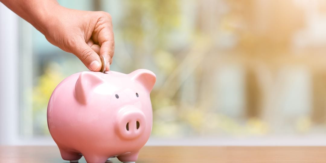 Mano de una personas insertando una moneda en un cochinito de porcelana rosa para su plan de ahorros, quiere comprar casa.