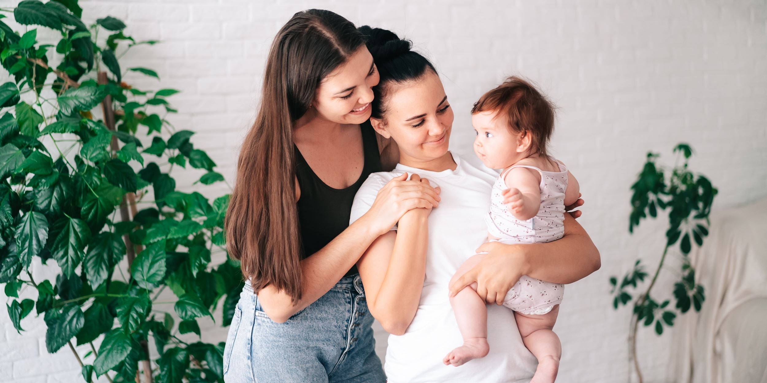 Crédito Rosa Infonavit. Pareja joven de mujeres con una bebé en brazos. Están dentro de su casa propia disfrutando el momento.