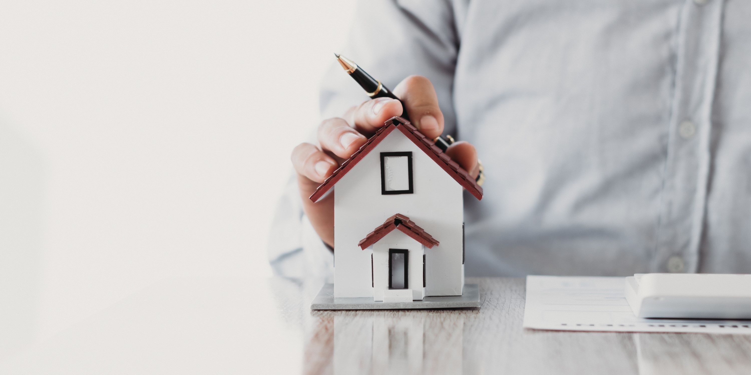 Hombre sujeta una casita en un escritorio. Tiene un bolígrafo, y papeles al lado. Representa la adquisición de un seguro de desempleo hipotecario.
