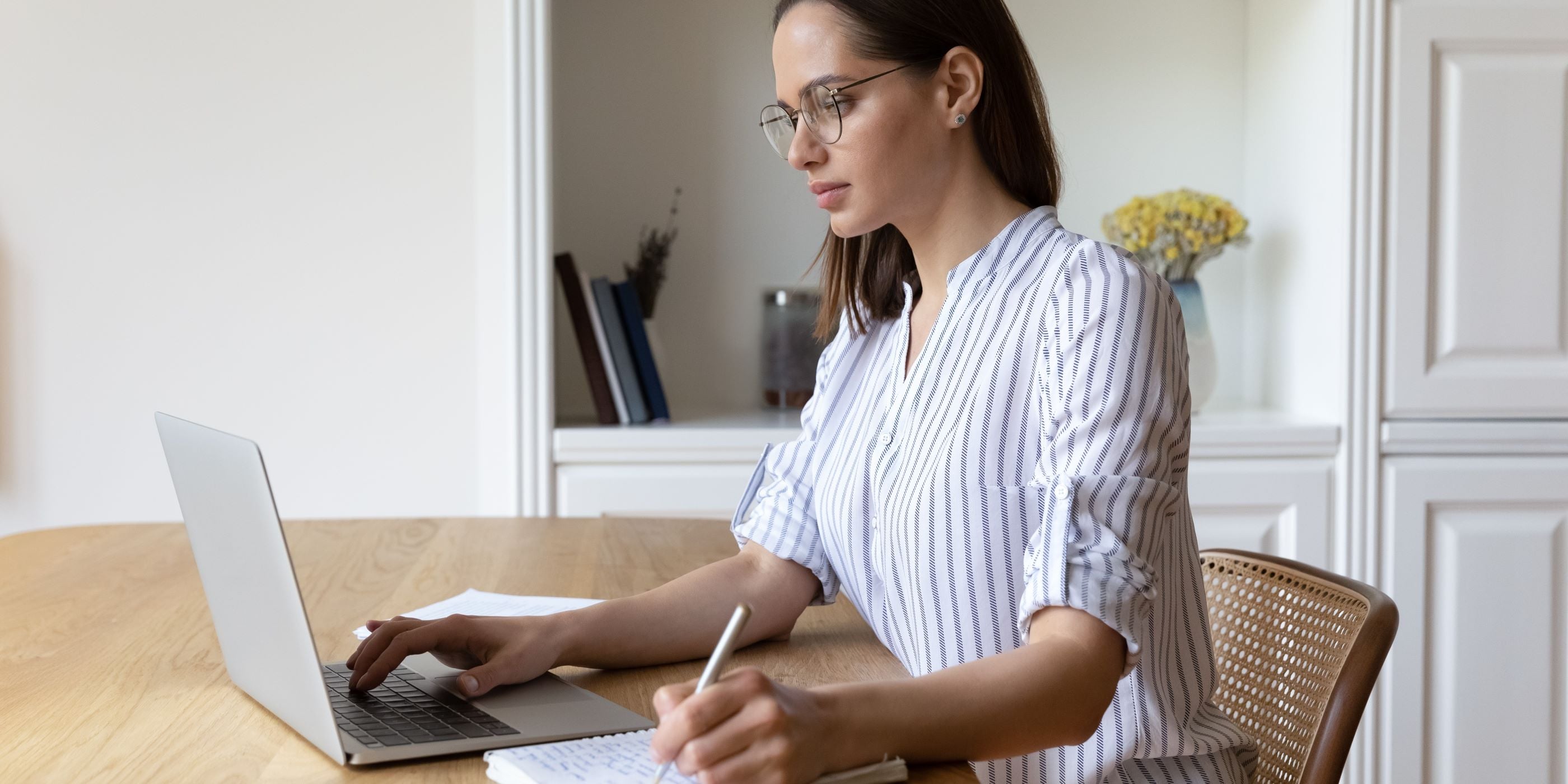 Mujer está sentada en su comedor; hace cálculos en su laptop, toma notas, y planea hacer inversiones inteligentes a futuro.