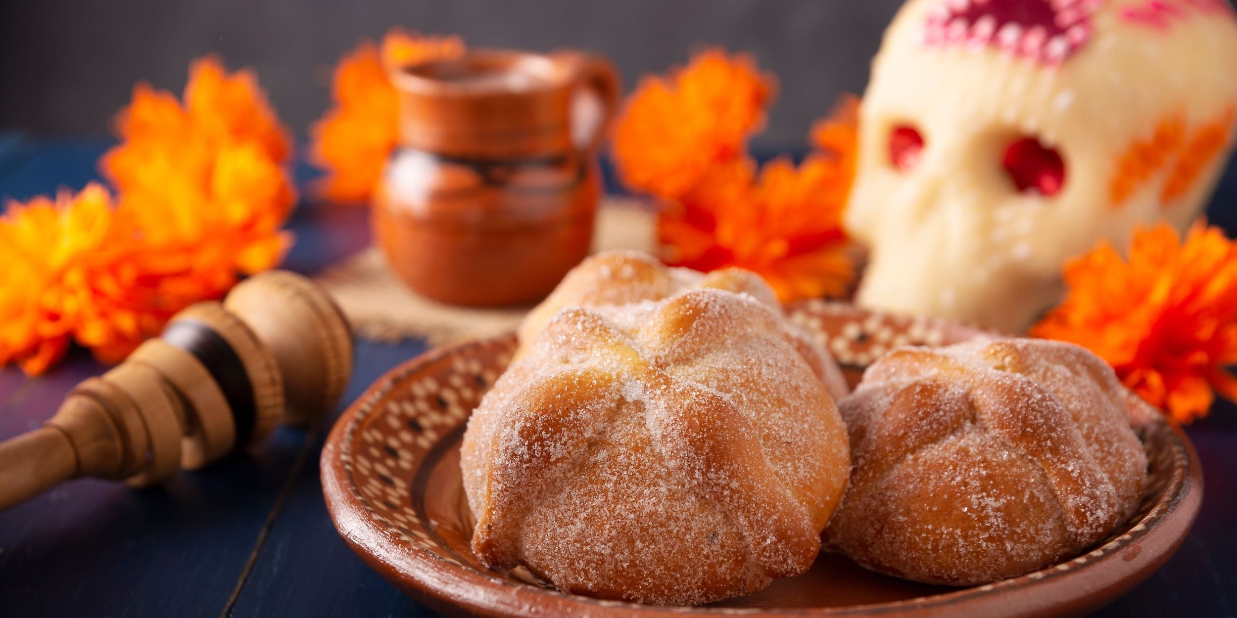 Pan de muerto, un molinillo de madera para hacer chocolate caliente, una calaverita de azúcar y una jarrita de barro.