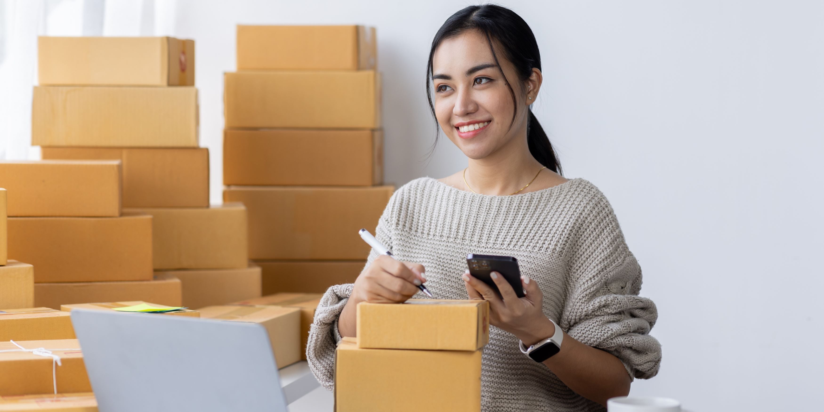 Mujer comienza su emprendimiento. Sostiene un celular y pluma frente a su computadora. Detrás tiene cajas con pedidos.