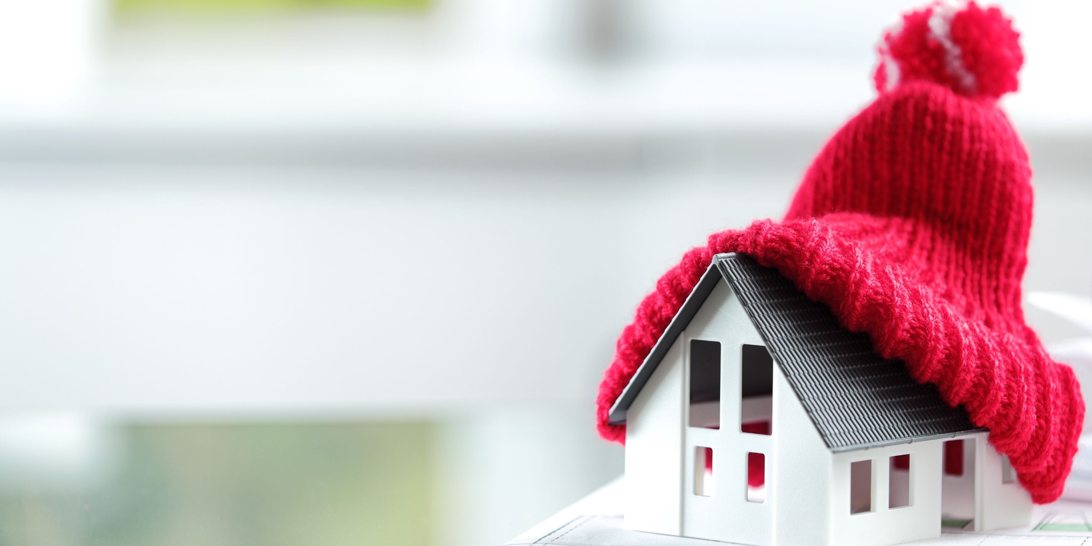 Casa miniatura en un escritorio. Tiene un gorro de estambre color rojo encima. Representa el cuidado del calor de una casa en tiempos de frío.