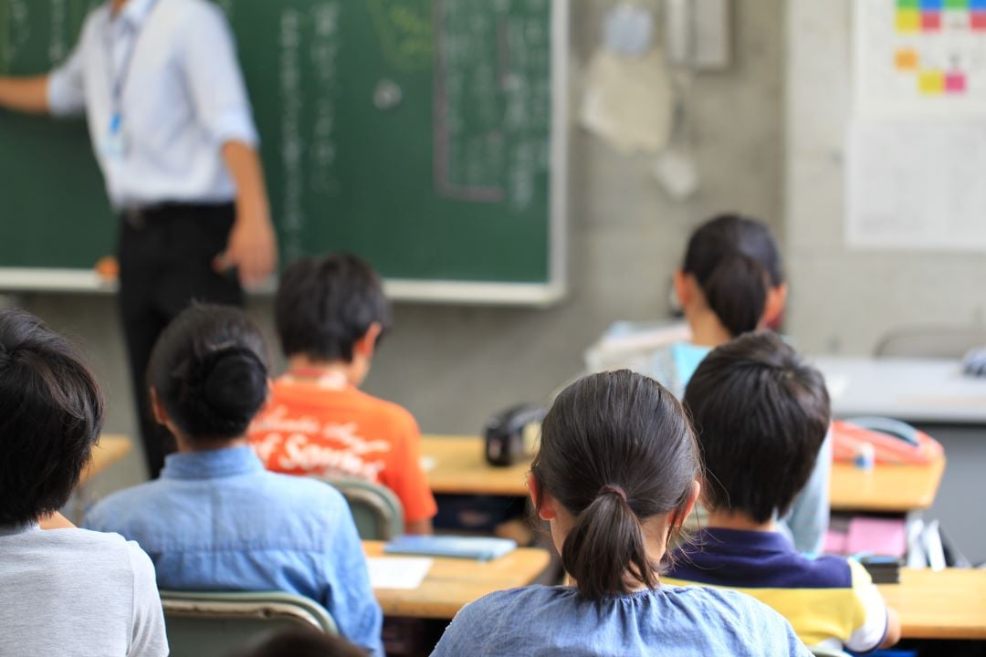 Niños en salón de clases. Hay un profesor al fondo, y niños tomando notas.