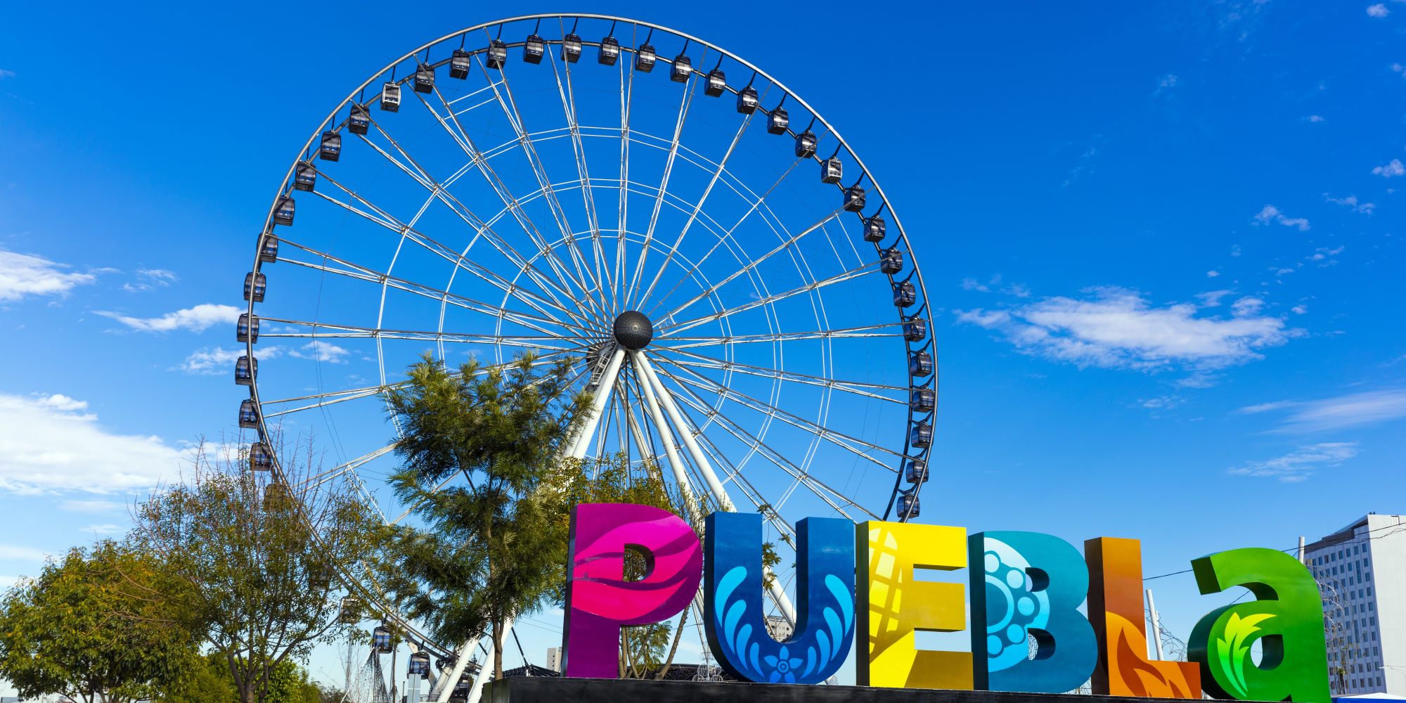 Explora los encantos turísticos de Puebla. Letras gigantes de PUEBLA están en primer plano, en segundo la gran rueda de la noria.